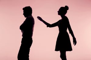 Image of boyfriend and girlfriend man turned other way with arms crossed distant and angry woman standing behind man with hand arm reached out for shoulder pink background black shadows outlines